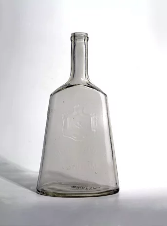 Przezroczysta szklana butelka. W podstawie ma owal. Dolna część lekko zwęża się ku górze i przypomina kształtem dzwonek. Zakończona jest u góry długą, prostą szyjką.