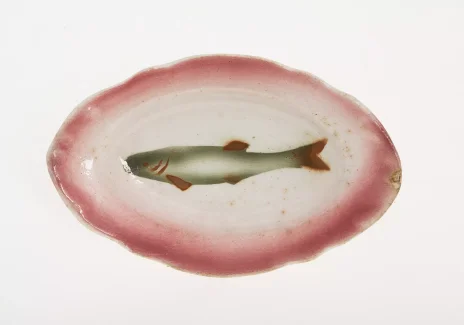 Owalny jasny talerz z nierównym brzegiem. Na środku namalowany podłużna ryba. Brzeg talerza i ryba w kolorze różowym.