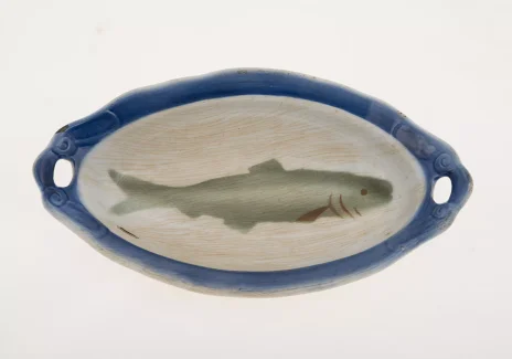 Podłużny talerz z niebieskim brzegiem i jasnym wnętrzem. Na środku namalowana podłużna ryba.