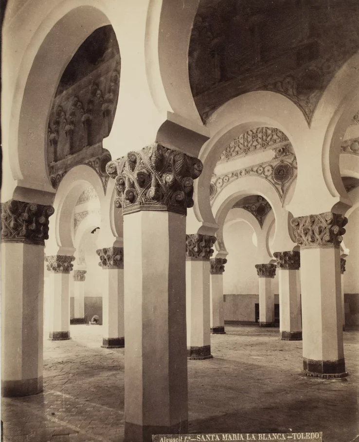 Fragment wnętrza synagogi orientalnej. Liczne białe sześciokątne kolumny. Ich zwieńczenie jest ciemne i dekoracyjne. Kolumny tworzą między sobą półkoliste łuki.