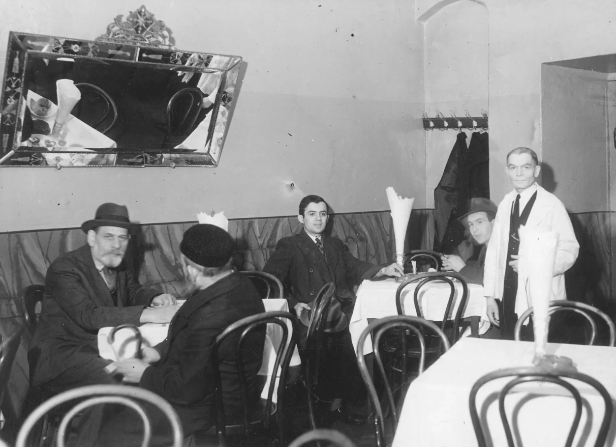 Czarno-biała fotografia wnętrza restauracji. Kilka stolików nakrytych białymi obrusami. Czworo gości przy stolikach. Kelner w bialym fartuchu. Na ścianie lustro.
