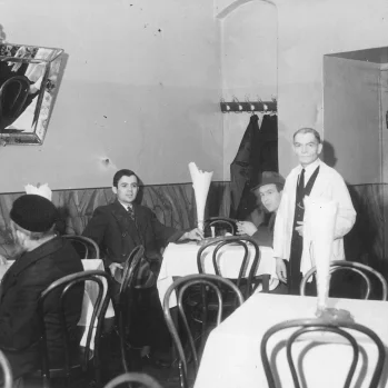 Czarno-biała fotografia wnętrza restauracji. Kilka stolików nakrytych białymi obrusami. Czworo gości przy stolikach. Kelner w bialym fartuchu. Na ścianie lustro.