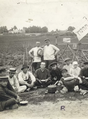 Czarno-biała fotografia. Grupa ludzi w roboczych ubraniach pozuje do zdjęcia na tle rozległego pola.