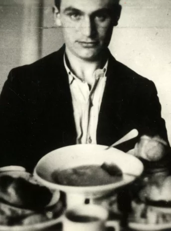 Czarno-biała fotografia młodego mężczyzny. Przed nim kilka talrzy wypelnionych jedzeniem.