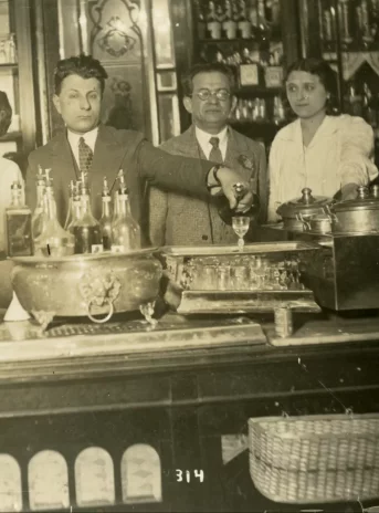 Fotografia w kolorze sepii przedstawia pięć osób stojących za bufetem. Trzy kobiety i dwóch mężczyzn. Na ladzie kasa, nalewak do piwa. Za nimi półki wypełnione butelkami.