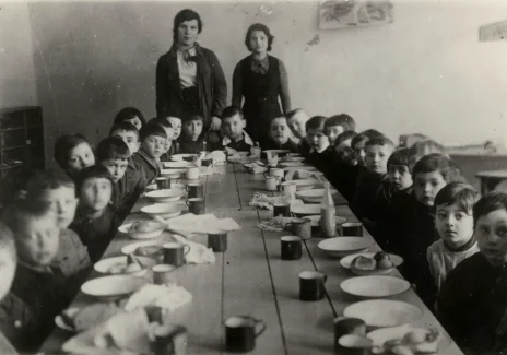 Czarno-biała fotografia wnętrza. Duża grupa małych dzieci siedzi ciasno przy stole. Przed nimi talerze i kubki. U szczytu stołu dwie dorosłe kobiety.