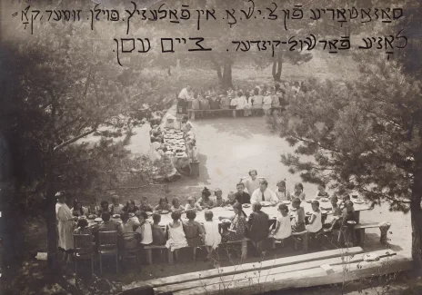 Czarno-biała fotografia parku lub ogrodu. Na trawie rozstawione długie stoły na około trzydzieści osób. Przy stołach, na ławkach, siedzą dzieci i kilka dorosłych osób. U góry zdjęcia napis w alfabecie hebrajskim.