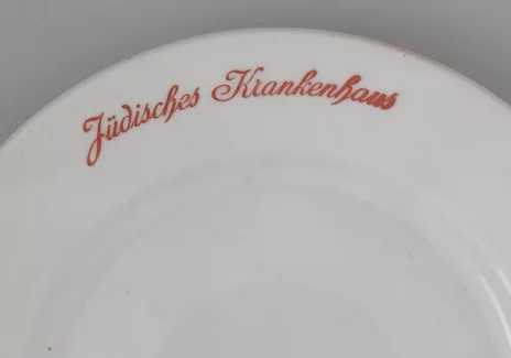 Dwa białe płaskie talerze. Na ich brzegach czarny napis w języku niemieckim: Jüdisches Krankenhaus, co się tłumaczy: Szpital żydowski.