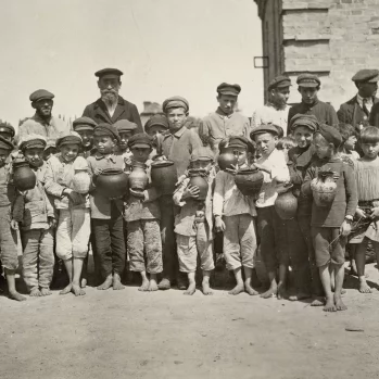 Czarno-biała fotografia przedstawia grupę ponad dwadzieściorga bosych dzieci. Za nimi kilku dorosłych. Stoją przed budynkiem, obok samochodu.