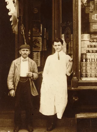 Fotografia w kolorze sepii. Dwóch mężczyzn przed sklepem, jeden w długim białym fartuchu. Nad sklepem napis w języku angielskim: Fine Delicatessen. M. Bloom.