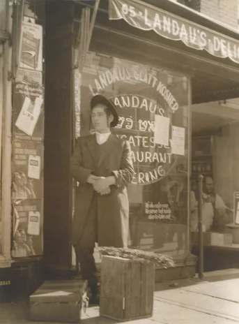 Fotografia w kolorze sepii. Mężczyzna w kapeluszu i płaszczu przed oszkloną witryną sklepu.