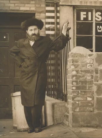 Fotografia w kolorze sepii. Brodaty mężczyzna w wysokiej czapce przed drzwiami do budynku. Na szybie obok napis po angielsku i hebrajsku: Fish Market, czyli Targ rybny.