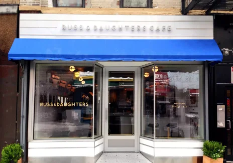 Kolorowa fotografia wejścia do kawiarni. Nad wejściem, nad niebieską markizą, napis: Russ & Daughters.
