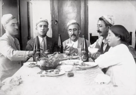 Czarno-biała fotografia. Czterech mężczyzn i kobieta przy zastawionym stole. Mężczyźni w żydowskich czapkach.