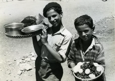 Czarno-biała fotografia. Dwóch chłopców niesie jedzenie na tacach.
