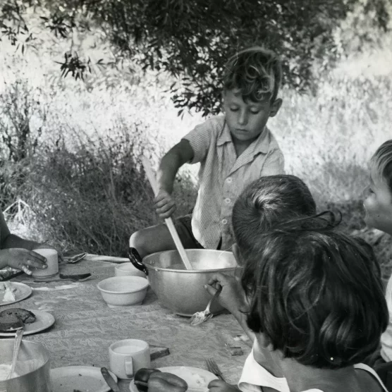 Czarno-biała fotografia. Siedmioro dzieci przy stole. Na nim talerze i naczycia. Jeden chłopiec nalewa zupę z miski.