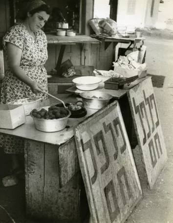 Czarno-biała fotografia.Stragan. Na straganie naczynia i jedzenie. Kobieta przekłada jedzenie dużą łyżką.
