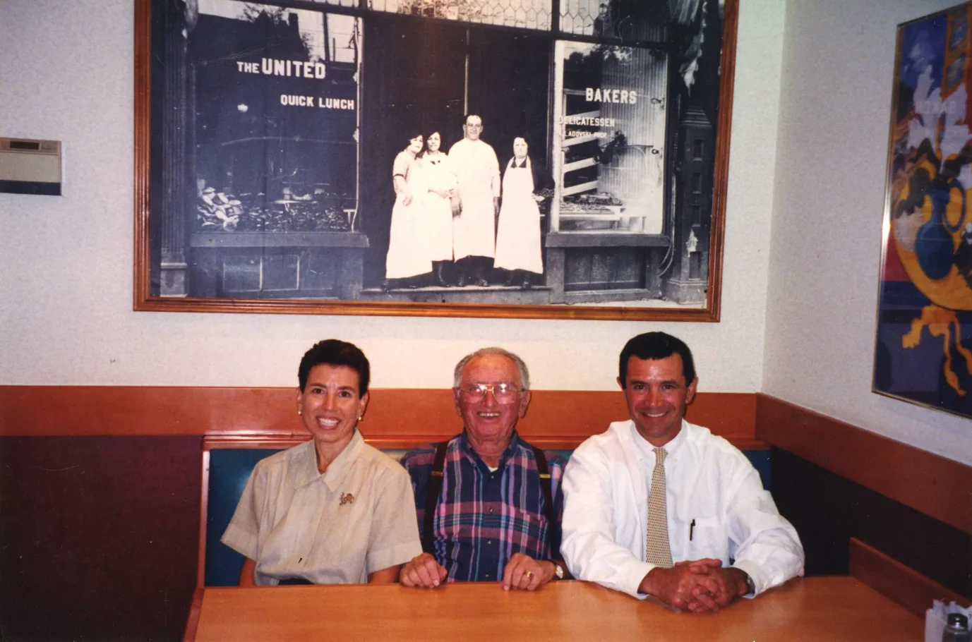 Kobieta i dwóch mężczyzn pozują do zdjęcia. Siedzą za stołem. Na ścianie za nimi duża czarno-biała fotografia.