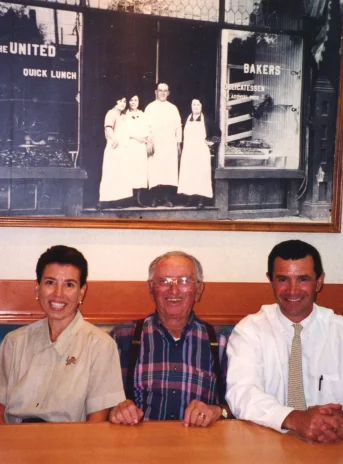 Kobieta i dwóch mężczyzn pozują do zdjęcia. Siedzą za stołem. Na ścianie za nimi duża czarno-biała fotografia.