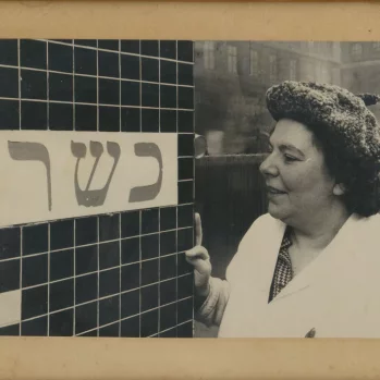 Kobieta w białym kostiumie, z gęstymi kręconymi ciemnymi włosami sto przy ścianie ułożonej z małych, czarnych kafelków, na której jest tabliczka z napisem w alfabecie hebrajskim.