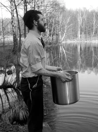 Black and white photography. A young man stands on the shore of a lake. In his hands he holds a large pot. In the background there is a forest.