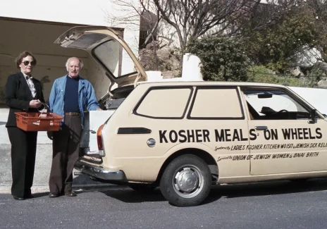 Two men are standing by the open trunk of a green car. The man on the left is dressed in black, has sunglasses, and holds a shopping cart in his hands. An older man on the right has a blue jacket. On the side of the car there is an inscription: Kosher meals on wheels, which means kosher food on wheels