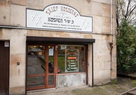 The front of the store, the gray walls of the glazed shop window and the entrance door. Above the door there is a sign with the Polish inscription Kosher Shop and text in the Hebrew alphabet.