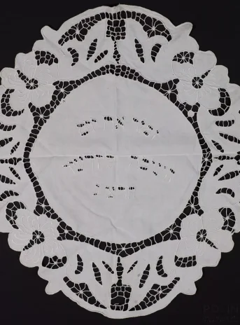 Biała serwetka w kształcie kwadratu z zaokrąglonymi rogami. Szeroki brzeg stanowi ażurowa, kwiatwa dekoracja. Na gładkim białym środku wyhaftowany tekst w alfabecie hebrajskim.