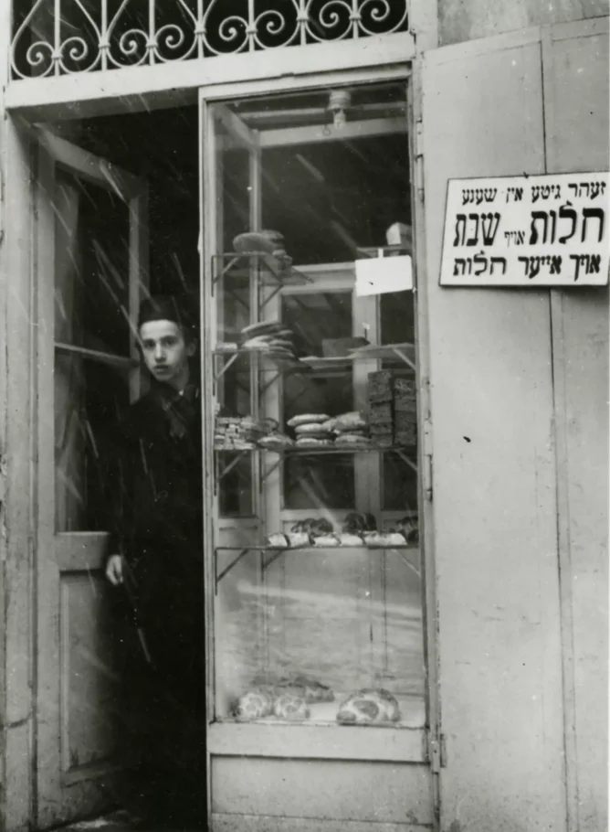 Black and white photography. Shop window. In the narrow door, a teenage boy. In the narrow window of the site shelf with bread.