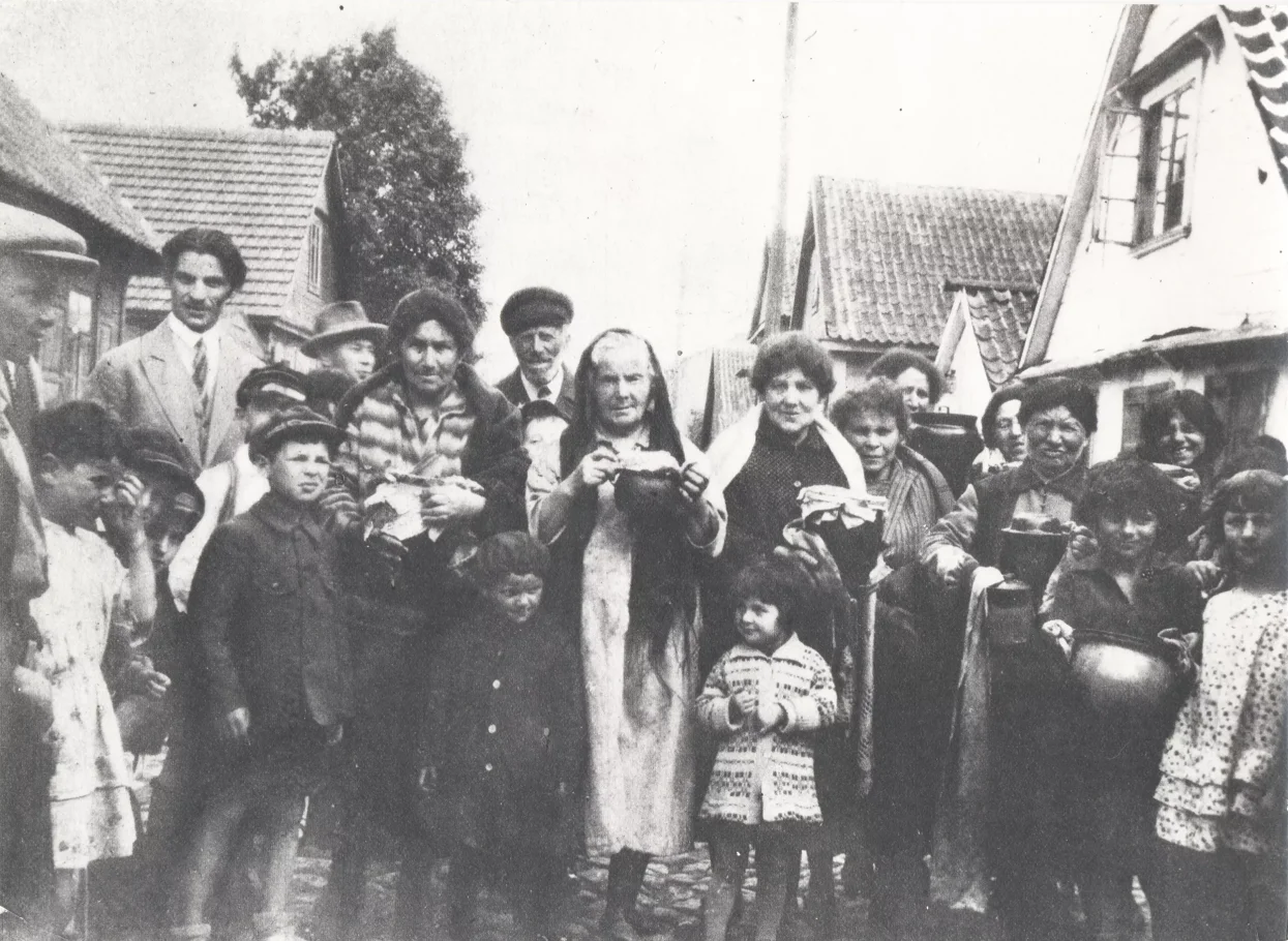 Black and white photo. Women, men and children pose for a photo. Females hold dishes in their hands. In the background, the roofs of houses.