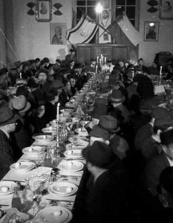 Black and white photo. 3 long set tables. Along them on both sides sit many people.
