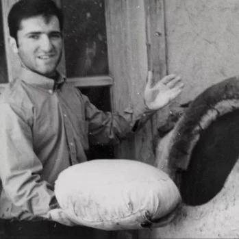 Black and white photography. A smiling young man poses by the bread oven. In his right hand he holds a dough wrapped in canvas. His left hand is slightly raised.