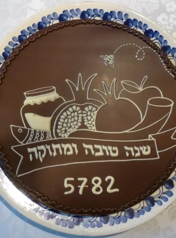 Photo of chocolate cake from above. Drawn a bright pattern depicting fish, vegetables, fruits and a jar of honey, below the inscription in the Hebrew alphabet and the number 5782.