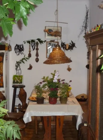 Kitchen interior. On the right side there is a large brown sideboard, next to the wall a small table covered with a white tablecloth with plants in pots placed on it. On the wall attached ornaments, Throughout the room a lot of plants.