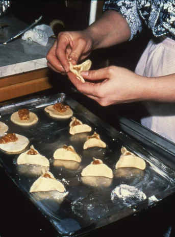 On the tray arranged in rows cookies with stuffing in the shape of a triangle. A woman forms one cake.