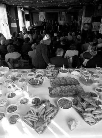 Black and white photo of a large room. In the foreground, a very large table covered with a white tablecloth. On the table stand various dishes. Behind the table, in the further part of the room sits a large group of people.