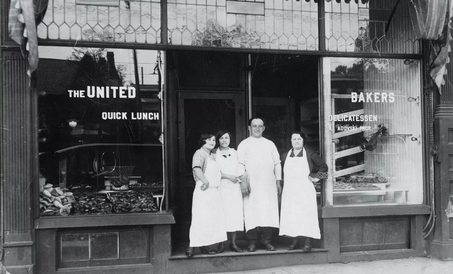 Czarno-biała fotogafia. Przeszklona witryna sklepowa. W drzwiach 3 kobiety i mężczyzna w długich białych fartuchch. Pozują do zdjęcia.