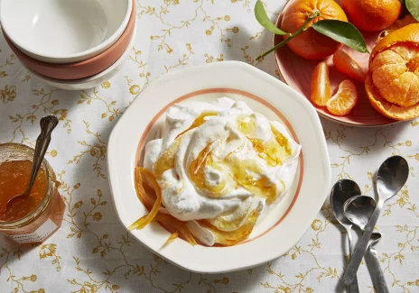 Biały talerz z białym kremem i fragmentami pomarańczowej skórki. Obok słoik z pomarańczowa konfiturą i talerz z mandarynkami.