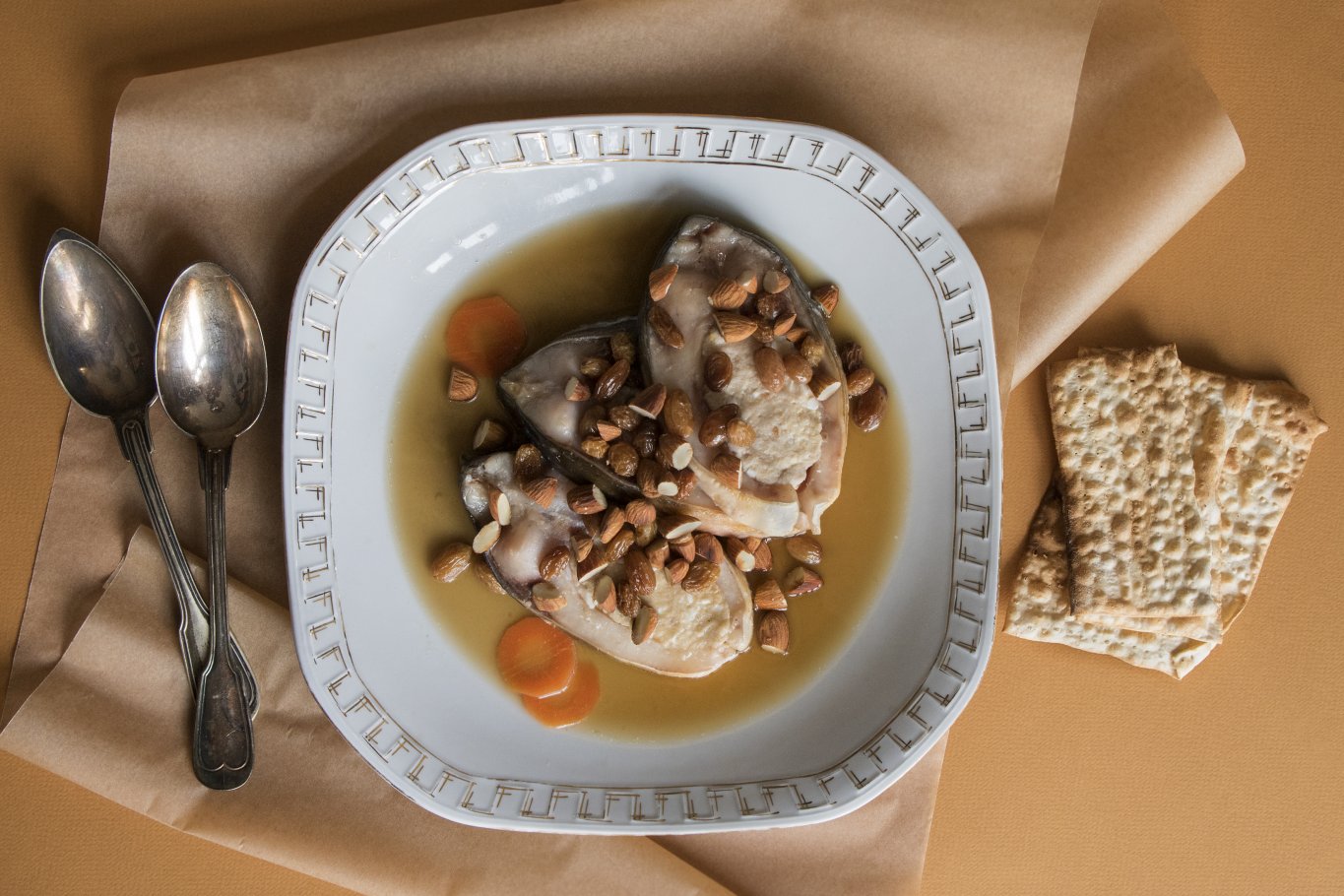 Głęboki, porcelanowy talerz wypełniony karpiem po żydowsku. Obok talerza leżą dwie ozdobne łyżki oraz maca.