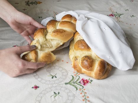 Dwie rumiane chałki owinięte w białą szmatkę. Kobiece dłonie urywają kawałek ciasta z jednej z nich.