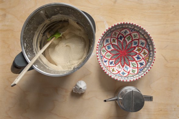 Kielich blendera, a w nim zmiksowany hummus. Obok leży miseczka z ozdobnym, orientalnym wzorem, głóka czosnku i metalowy imbryk.