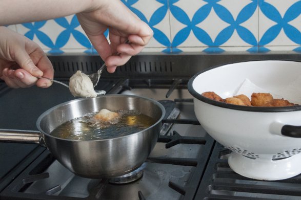 Osoba przygotowująca pączki umieszcza masę z ciasta w rozgrzanym oleju. Robi to za pomocą dwóch łyżek. Obok znajduje się biała waza, w której leżą usmażone pączki.