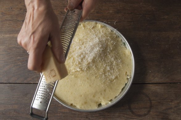 Osoba przygotowująca posiłek uciera parmezan na tarce. Robi to nad okrągłą formą wypełnioną składnikami na mina de espinaca. Starty parmezan ląduje na masę ziemniaczaną.