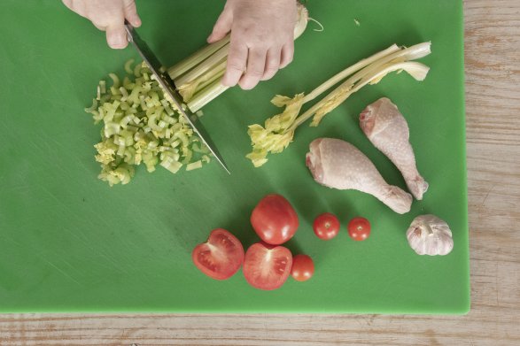 Na zielonej desce do krojenia osoba przygotowująca posiłek odcina zielone liście selera od korzenia. Obok selera leżą dwie pałki z kurczaka, pomidory i główka czosnku.