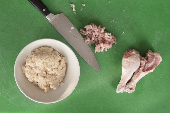 Na zielonej desce ułożone są dwie pałki kurczaka, kupka mięsa mielonego, ostry nóż oraz miseczka z jasną pulpą.