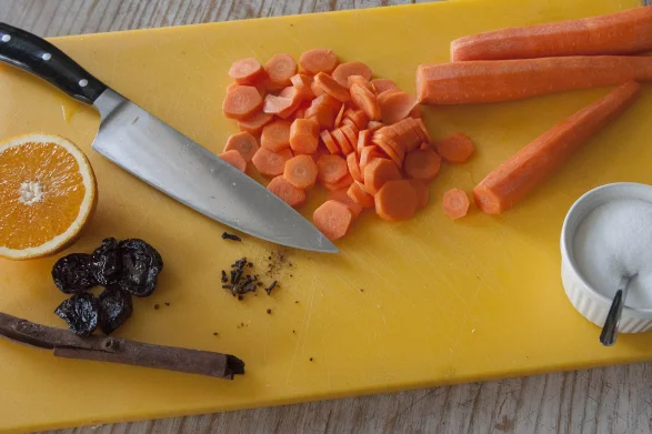 Na żółtej desce do krojenia rozłożone są pokrojone marchewki, pół pomarańczy, śliwki, laska cynamonu i cukier w porcelanowym pojemniczku. Obok jest odłożony nóż.
