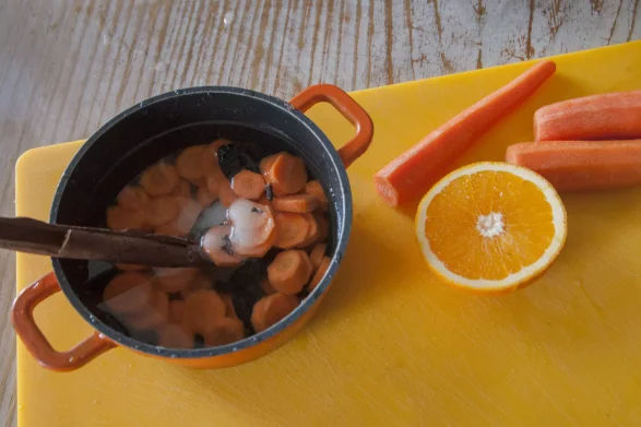 Na żółtej desce leży garnek wypełniony wodą z pokrojonymi marchewkami i cynamonem. Obok znajdują się obrane marchewki i pół pomarańczy.