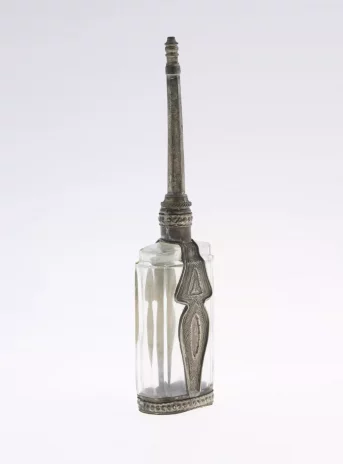 Naczynie na oliwę w formie prostokątnej butelki szklanej z metalowymi zdobieniami oraz długim metalowym lejkiem. Zdobiony ornamentami.