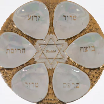 Round plate in shimmering golden color. In the middle is the pearl star of David. Between the star and the edge evenly distributed 6 pearl petals. The petals form with the star the shape of a flower. On each petal there is an inscription in the Hebrew alphabet.