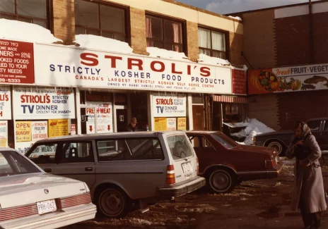 The front of the store. On a white sign above the door, the name of the store. On the walls and sites information and advertising slogans. Several cars are parked in front of the building.